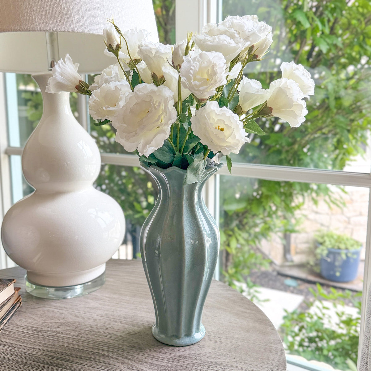 White flowers in a blue fluted vase on a table with a white lamp and window in the background