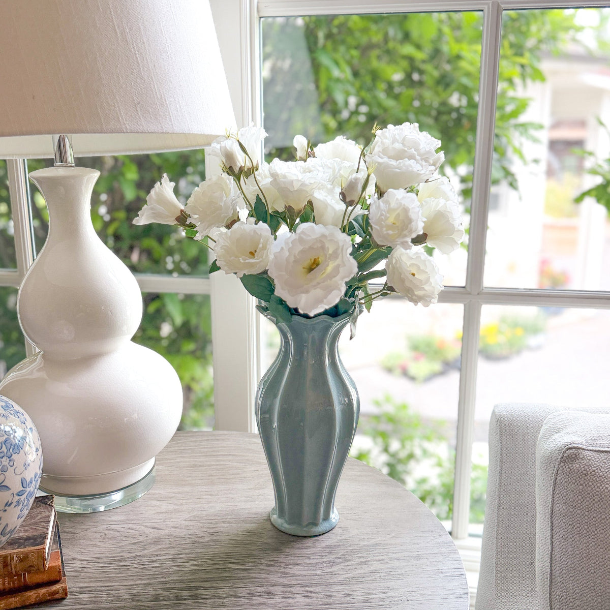 White flowers in a blue fluted vase on a table with a white lamp and window in the background