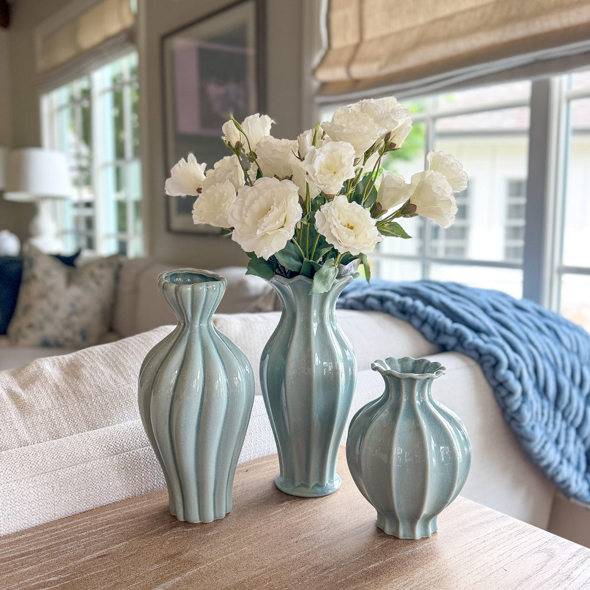 Three light blue fluted ceramic vases with white flowers on a wooden table in a living room.