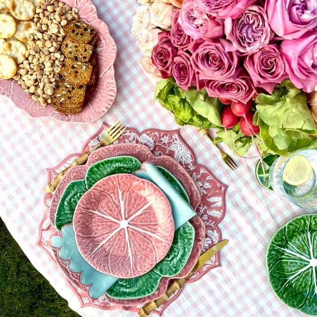 Table setting with pink and green plates, flowers, and food on a checkered tablecloth.