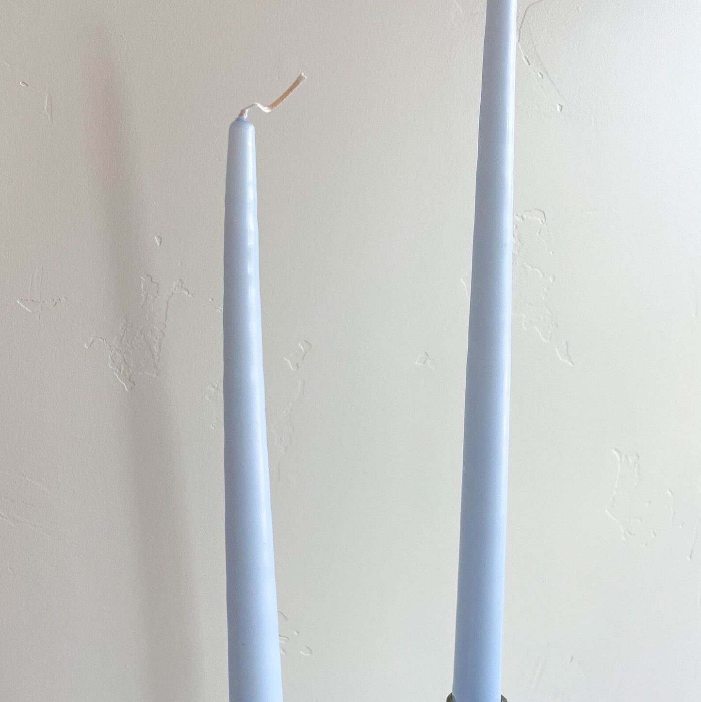 Two ice blue taper candles placed back to back on a surface, with a white wall in the background.