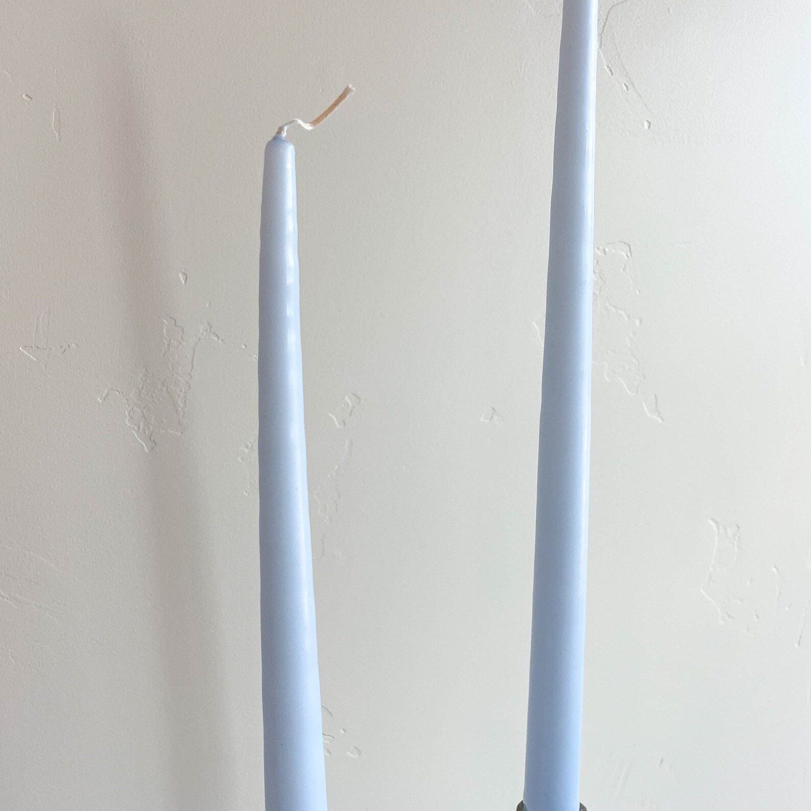 Two ice blue taper candles placed back to back on a surface, with a white wall in the background.