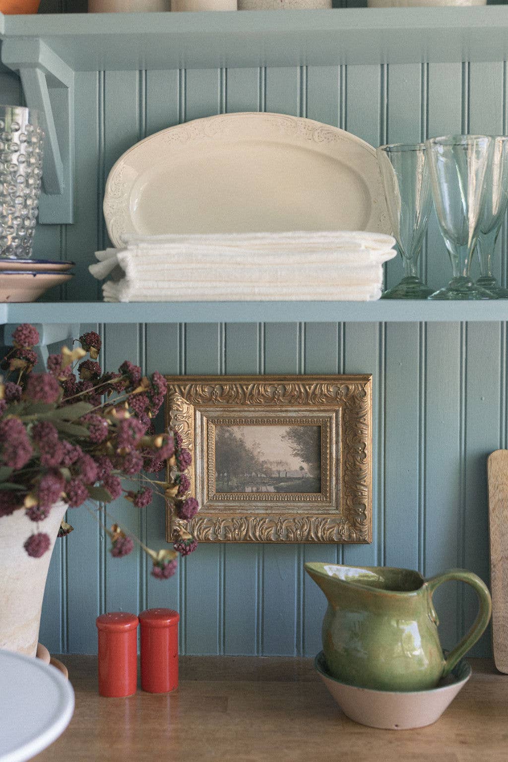 Decorative shelf with plates, a pitcher, and a framed picture against a blue paneled wall.